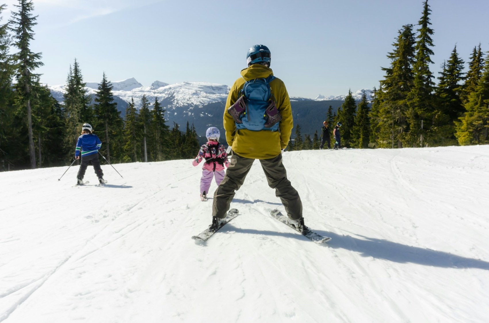 A parent and two children skiing a wide groomed run with evergreen forest behind