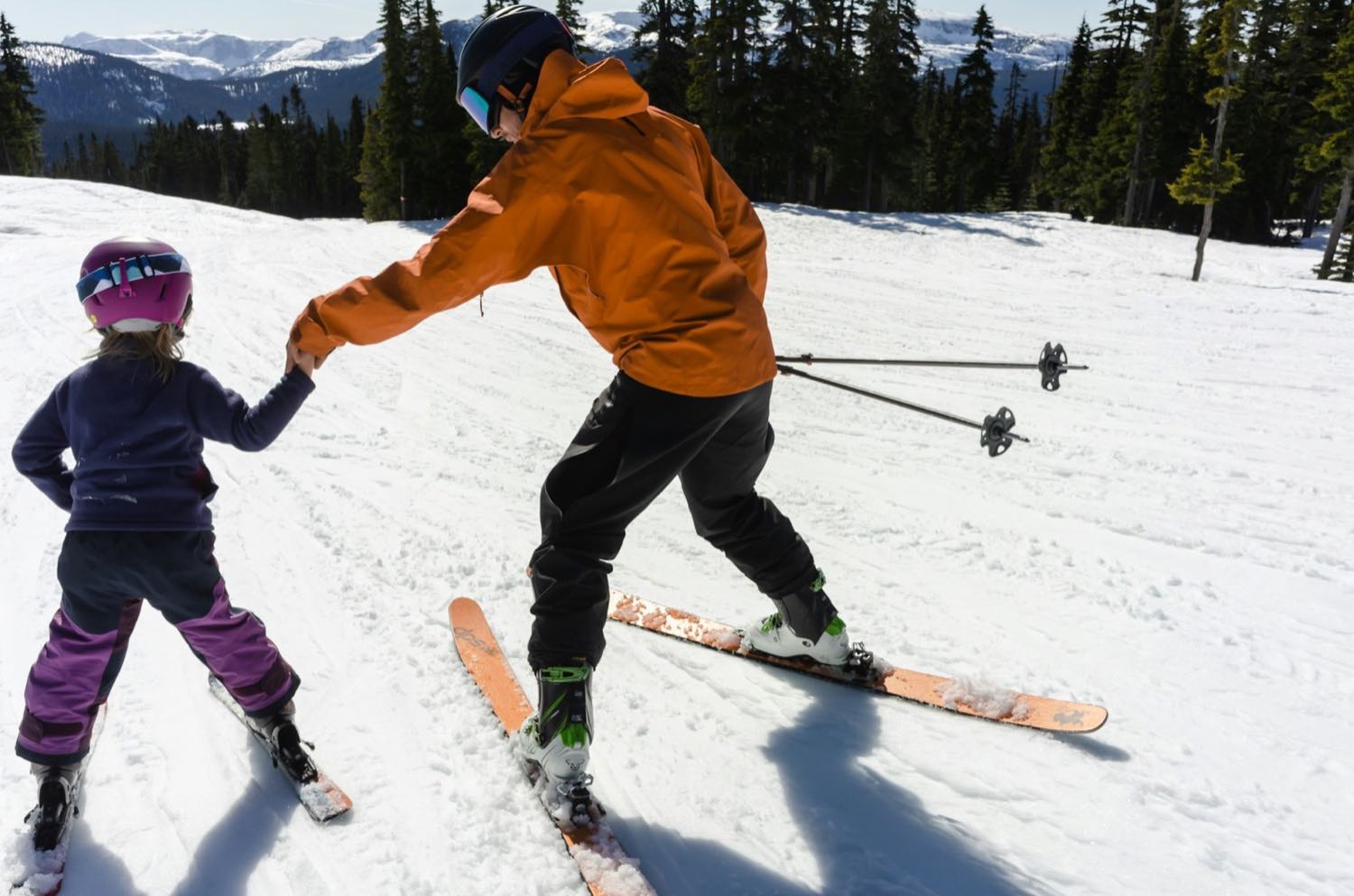 An adult holding a small child's hand as she learns to ski on a gentle slope