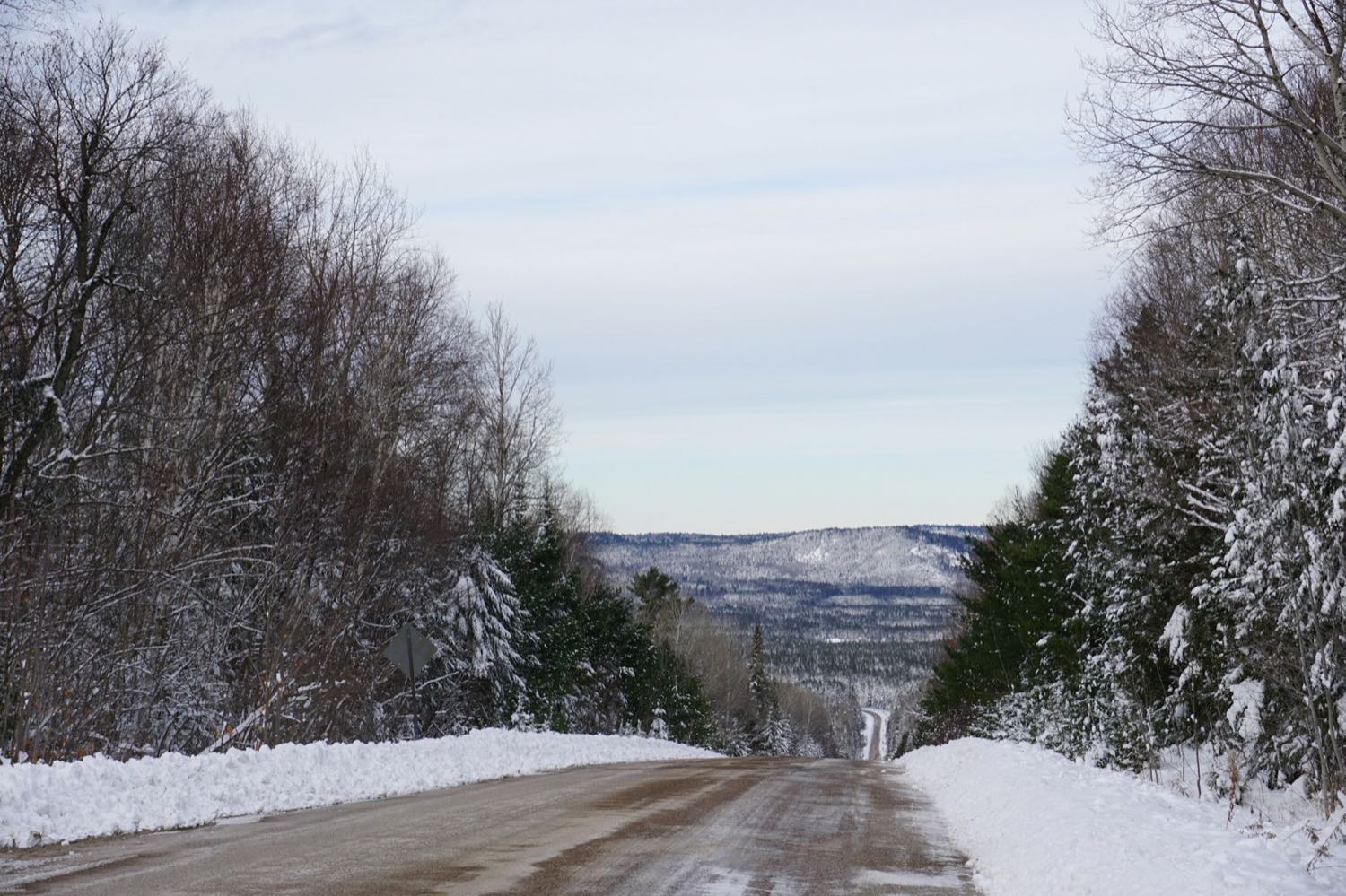 A snow-banked two-lane road running through mixed winter forest towards low hills