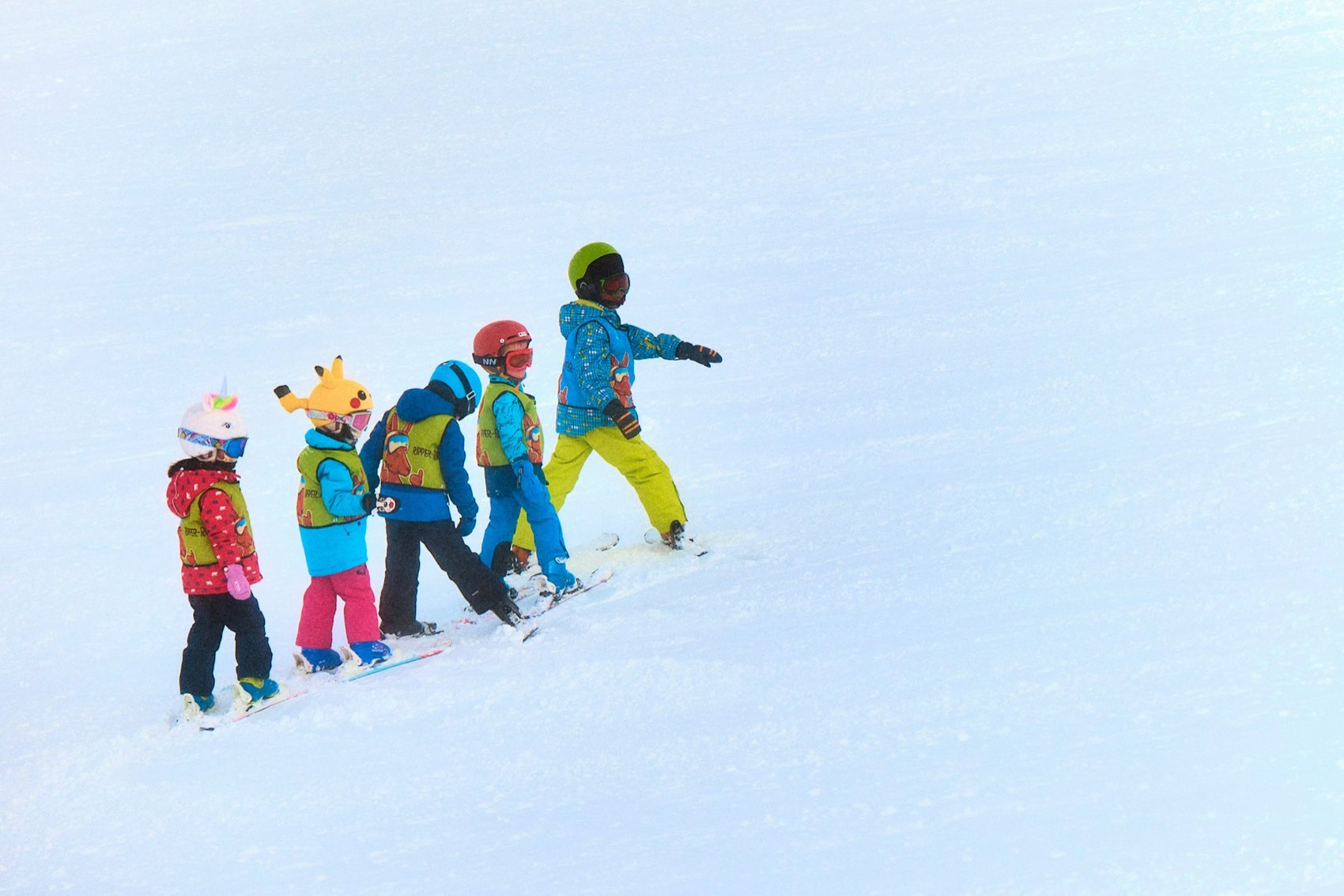 Five small children in colourful helmets and bibs walking in a line across the snow